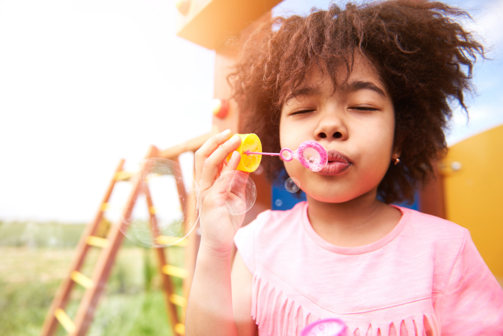 A child in a pink shirt blowing bubbles through a wand while standing in front of a playground.