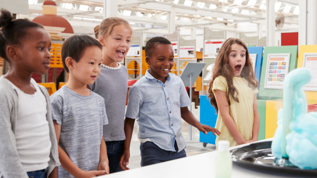Five kids react excitedly to green foam erupting from a beaker during a chemistry demonstration at a museum.