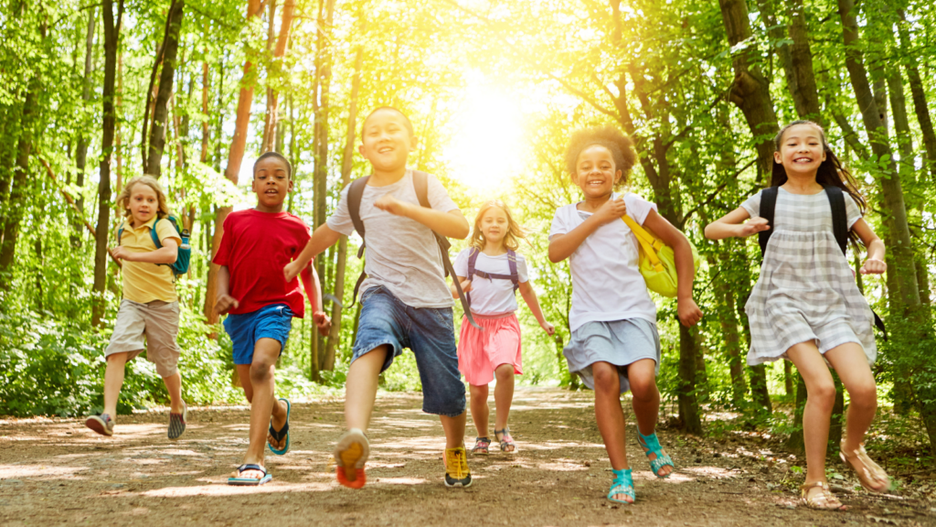 Six kids in t-shirts and shorts and wearing backpacks laugh and run towards the camera as the sun shines between trees behind them.