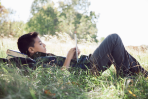 A boy uses a tablet while lying in a grassy field with his head on textbooks in his open backpack.
