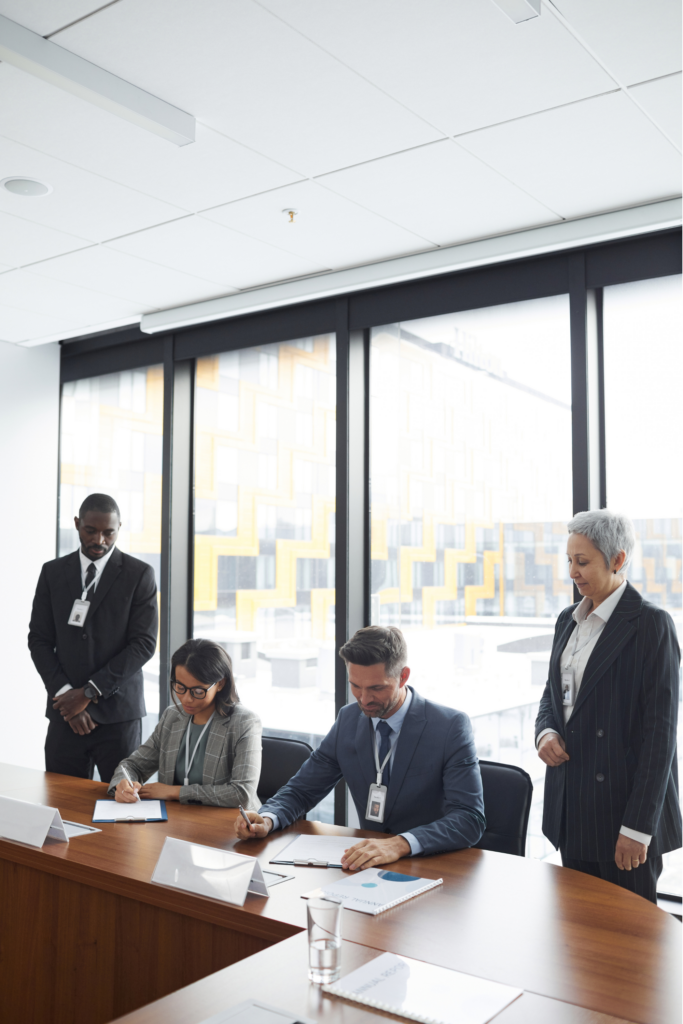 Two government officials wearing suits and nametags on lanyards sit at a conference table reviewing printed materials. Two aides, also wearing suits and nametags on lanyards, stand to either side of them.