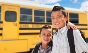 Two boys wearing backpacks pose for a photo in front of a school bus.