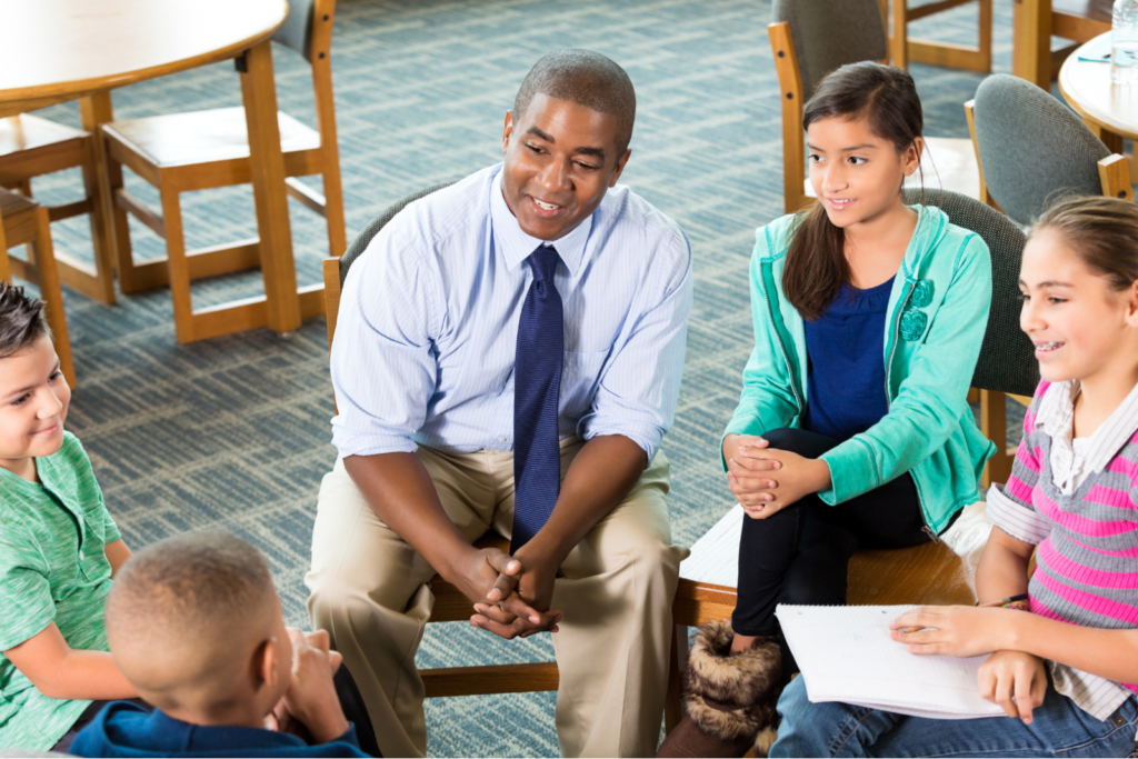 A group of middle school students and a teacher sit in a circle in the library and have a conversation.