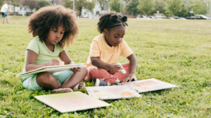 Two girls sit cross-legged in a grassy park doing crafts on large square paper.