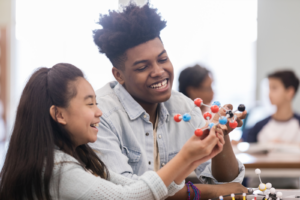 A high school student sits with a younger child in a library and is tutoring them in chemistry with a model of a molecule.