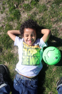 A curly-haired kid smiles and lies on the grass with his arms folded behind his head next to a bright green soccer ball.