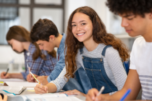 Four high school students sit at a long table taking notes from books in front of them.