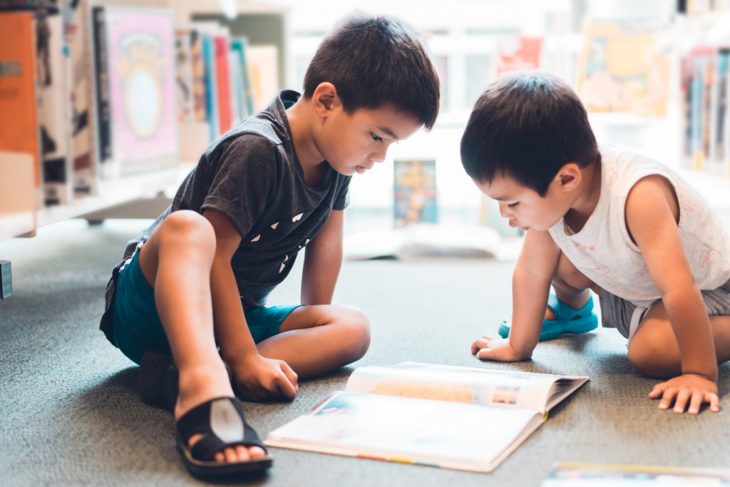 Two boys wearing shorts, short-sleeved shirts, and sandals sit on a carpet in a library and read an open picture book.