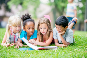 Four kids read a picture book together while lying on their stomachs in the grass.