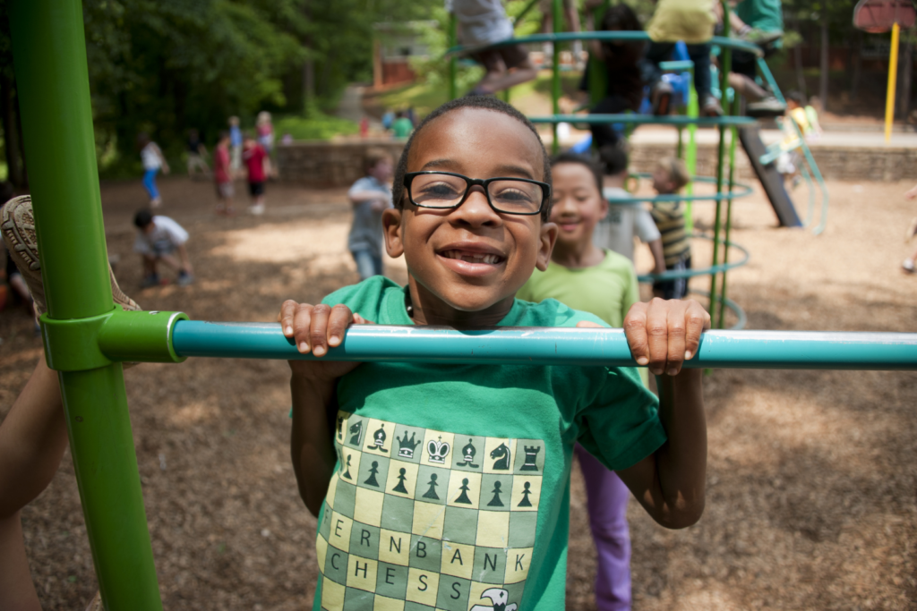 A boy smiles at the camera while holding onto a monkey bar on a playground.